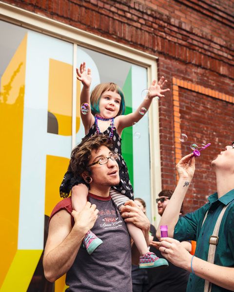 a parent carries their child on their shoulders while the other parent blows bubbles toward them