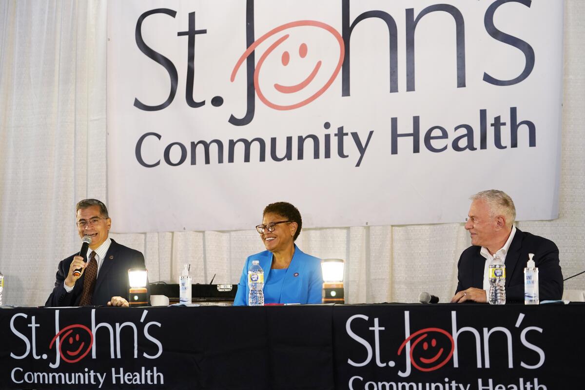 A man in dark suit and tie speaks into a mic while seated next to a woman in a blue suit and another man in a dark jacket