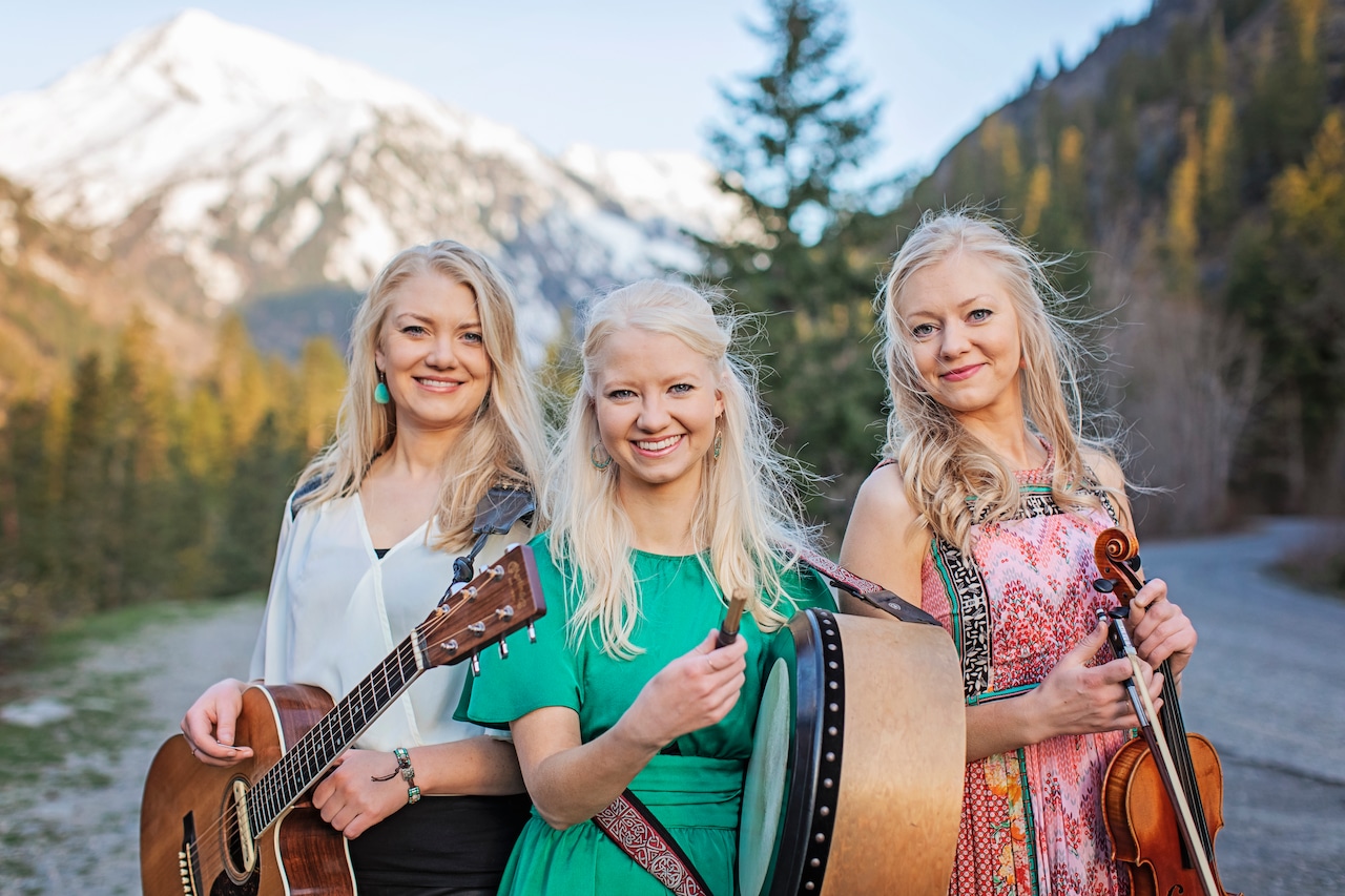 Three women hold instruments outdoors in front of a snow-capped mountain