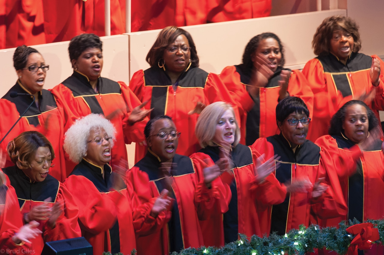 11 women wearing red choir robes with black trim stand in two rows, singing.
