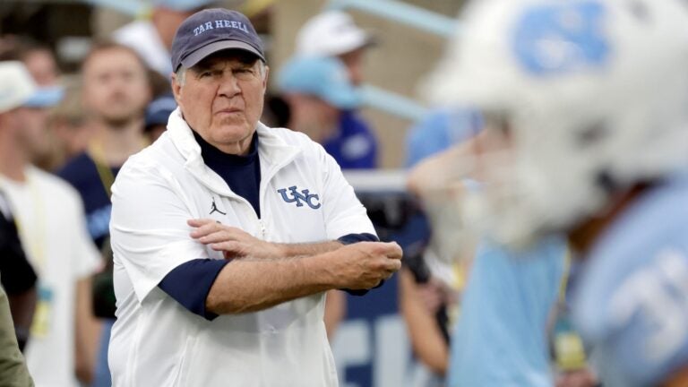 Bill Belichick watches his team warm up on the field before a game, wearing a navy "Tar Heels" ball cap and a white UNC quarter zip.