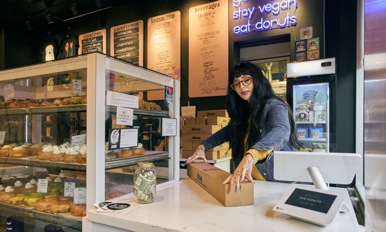 a woman at a counter closes the lid on a box of doughnuts at the counter of a bakery behind a glass cabinet full of doughnuts and desserts