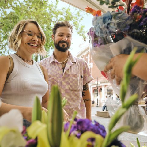 people smiling on a sunny day in front of bunches of flowers while someone off screen is handing them a bundle of cut flowers in a plastic bag