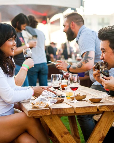 two people eating food and drinking wine at a picnic table during a busy food festival