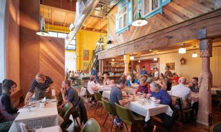 tables full of diners enjoy a meal in a lively, warmly lit restaurant with a bar in the background