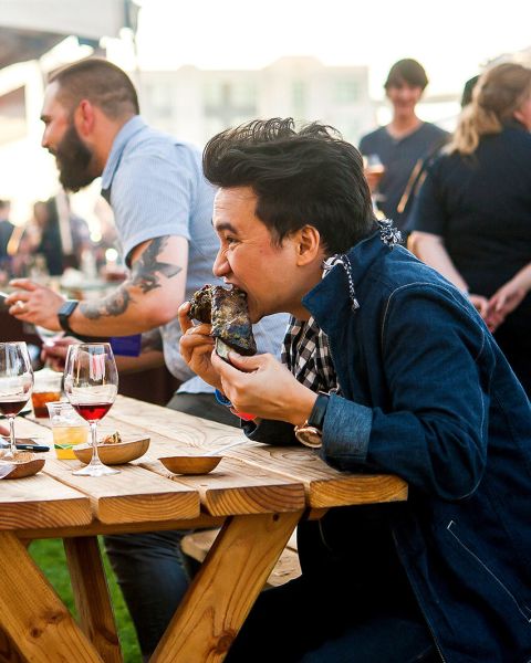 two people eating food and drinking wine at a picnic table during a busy food festival