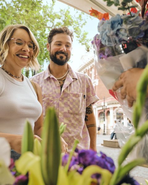 people smiling on a sunny day in front of bunches of flowers while someone off screen is handing them a bundle of cut flowers in a plastic bag