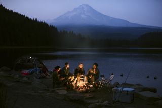 four people illuminated by campfire at a dimly lit campsite, backed by a serene lake and a large mountain obscured by mist
