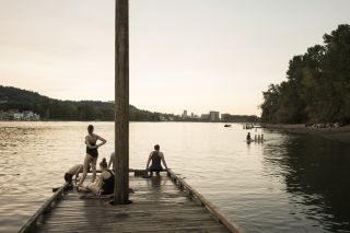 a wooden dock in a river — with swimmers standing, sitting or climbing out of the water — looks toward a city skyline in the fading sunlight