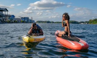 two women on paddleboards in a river with houseboats in the distance
