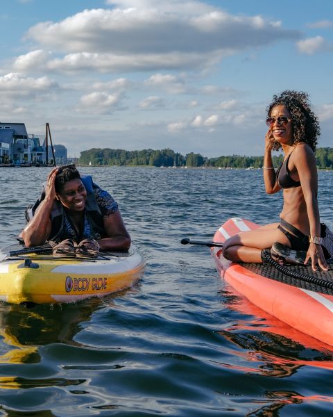 two women on paddleboards in a river with houseboats in the distance