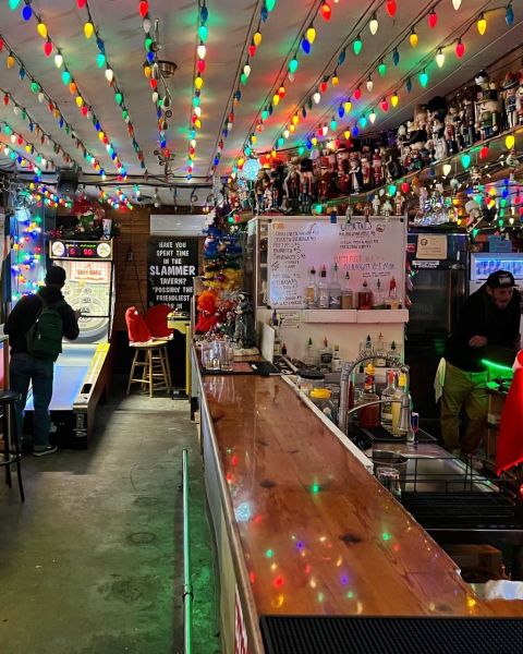 a patron plays Skee-Ball and a bartender leans against the backbar in a tavern lighted with Christmas lights