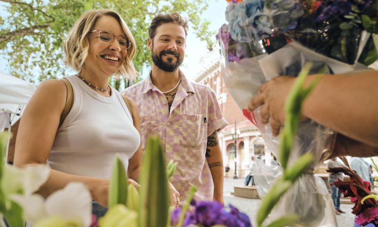 people smiling on a sunny day in front of bunches of flowers while someone off screen is handing them a bundle of cut flowers in a plastic bag