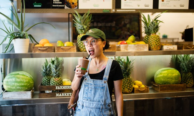 A woman sips a smoothie through a straw from a plastic cup in front of a display of fresh fruit