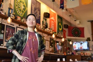 A woman in a plaid button-down and red t-shirt sits at a bar in front of a wall of women's sports memorabilia.