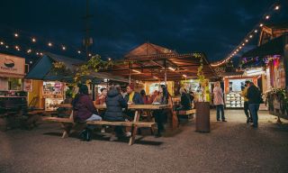 people seated at picnic tables talk and eat with food carts in the background