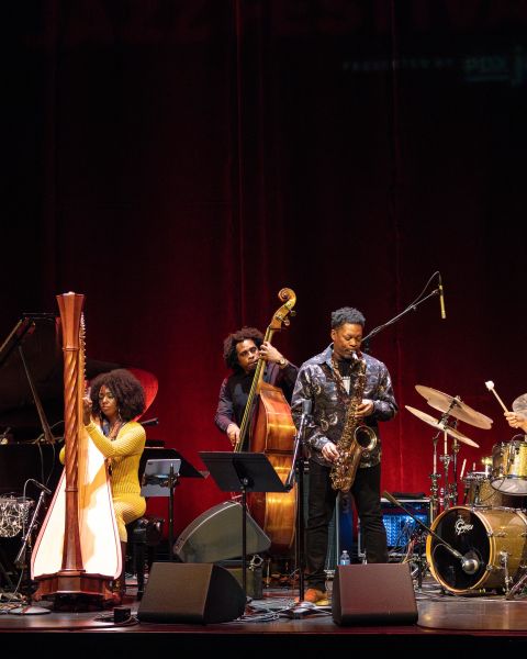 A band of five Jazz musicians performs on stage in front of a red curtain.