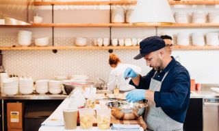 In a brightly lit kitchen, a man wearing , blue gloves, an apron and a baseball cap mixes ingredients inside a metallic bowl.