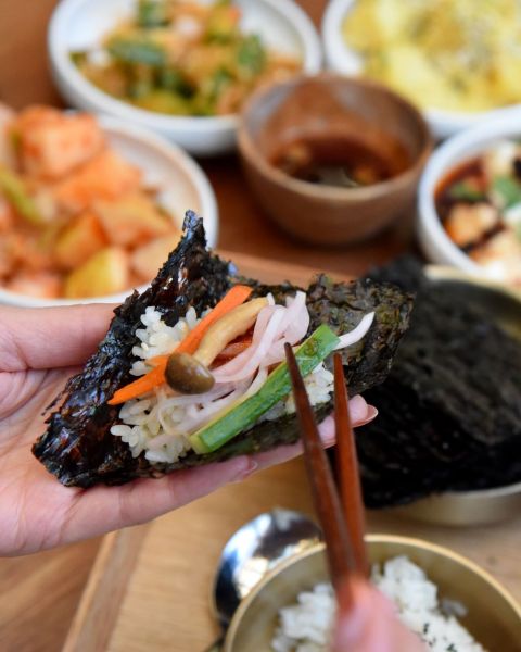 chopsticks reach for colorful vegetables atop a sheet of seaweed with a spread of Korean food in the background