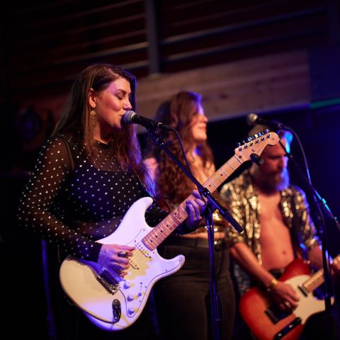 person sings in to a microphone while playing a guitar with two band members behind on a stage.