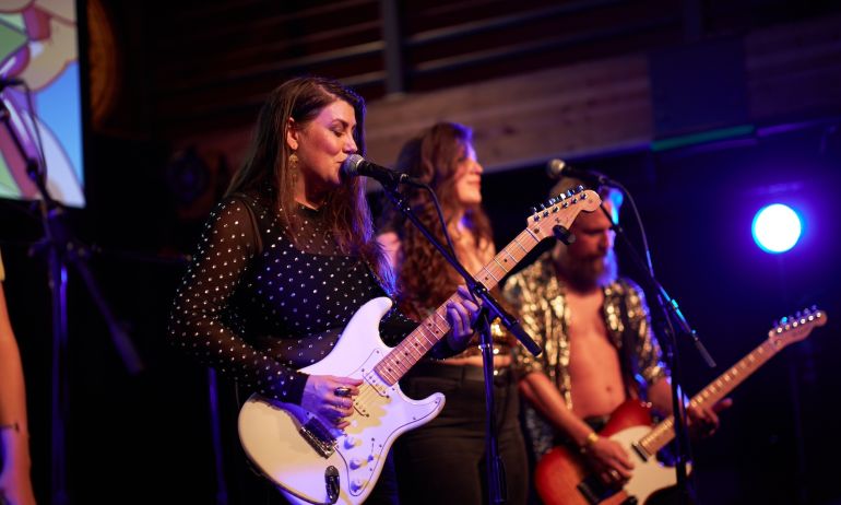 person sings in to a microphone while playing a guitar with two band members behind on a stage.