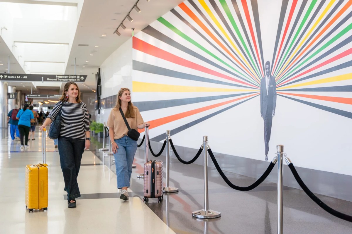 39 July events to experience—festivals, firework shows + more 2 Two travelers with luggage walk past a large colorful mural featuring a suited figure at Birmingham-Shuttlesworth International Airport
