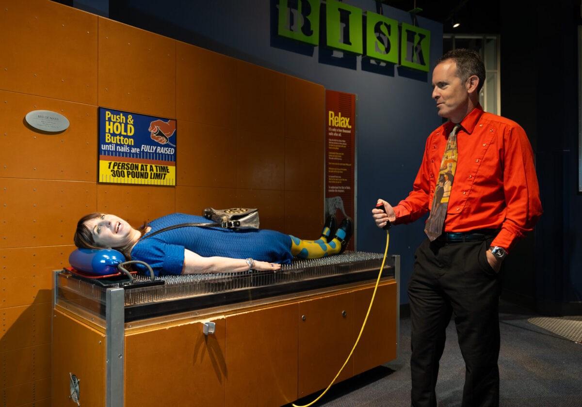 A woman lies smiling on the Bed of Nails exhibit at McWane Science Center while a man holds the activation cord beside her