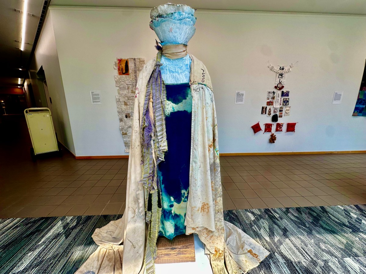 A sculptural dress installation featuring blue-dyed fabric and a flowing white robe with mixed materials, displayed in the Embodied exhibit gallery at Birmingham Public Library