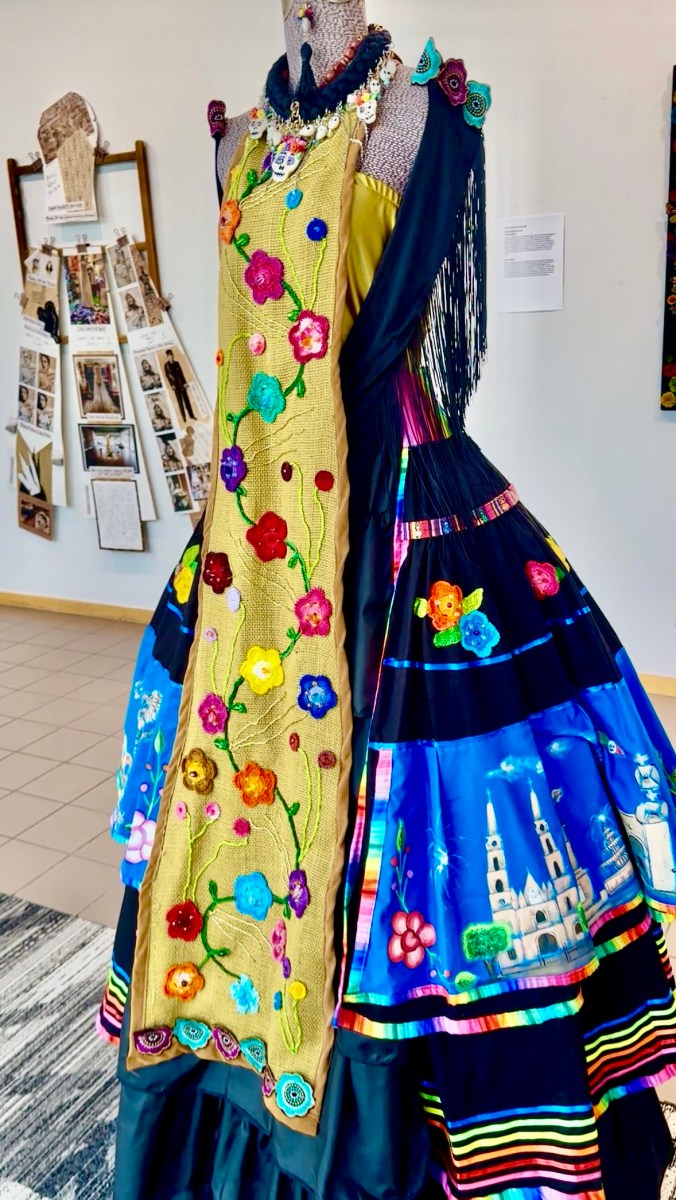 A dress on display featuring colorful embroidered flowers on burlap, a black fringed shawl, and a painted skirt depicting a cathedral, part of the Embodied exhibit at Birmingham Public Library