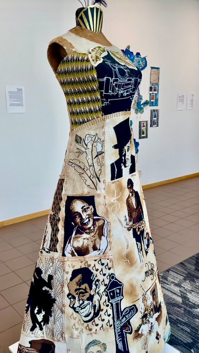 A dress on a mannequin featuring embroidered and printed portraits of jazz musicians, florals, and a Lenox Ave street sign, displayed in the gallery
