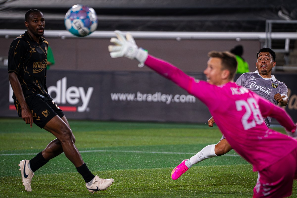 A goalkeeper in pink stretches to make a save during a soccer match as two outfield players look on