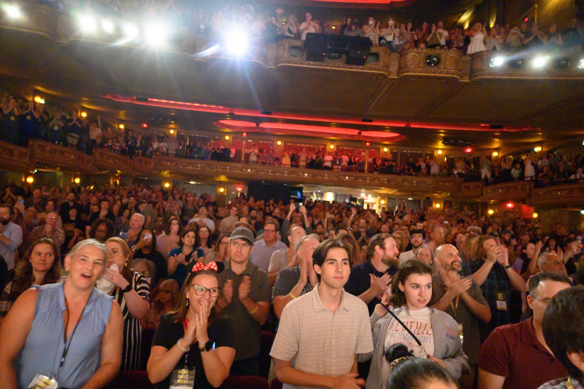 A packed theater audience gives a standing ovation, with multiple balcony levels filled with applauding and cheering attendees