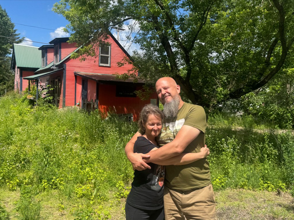 A woman and a man hug. There is an orange home behind them and a field of green plants.