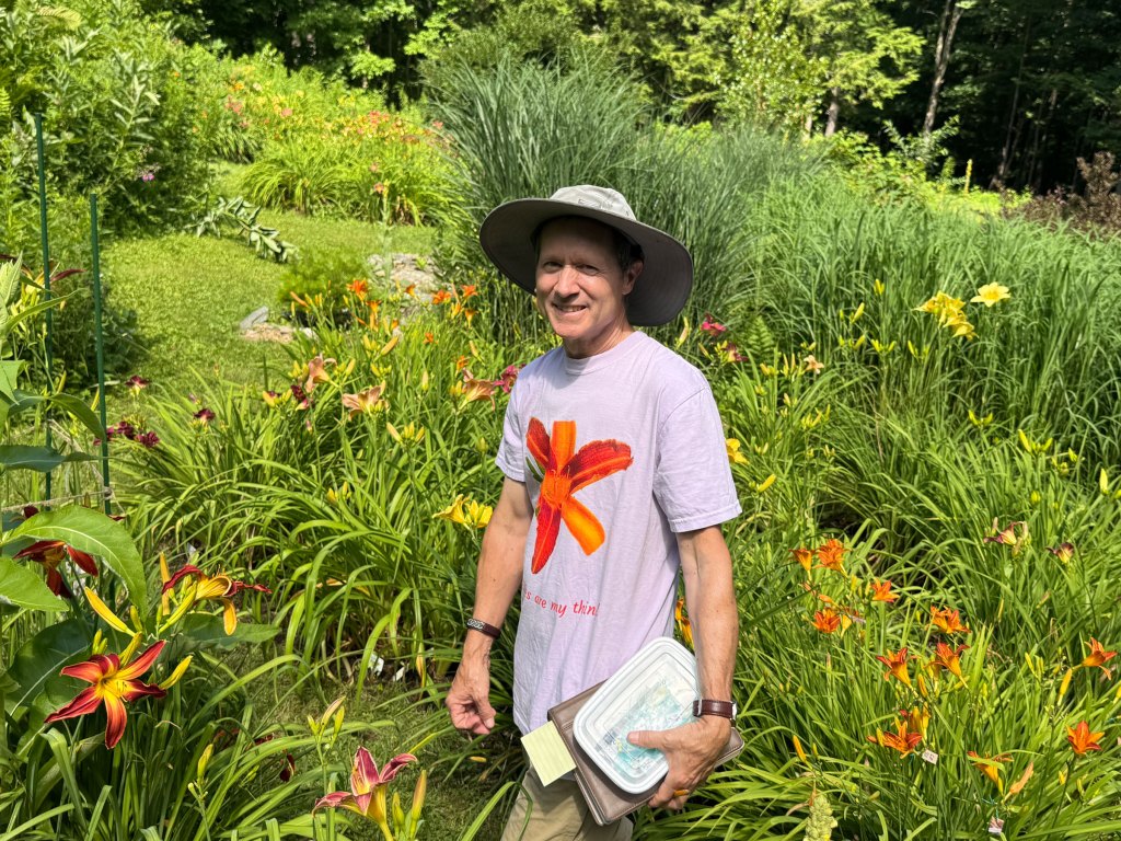 A man wearing a shirt with a daylily image on it stands in a colorful garden