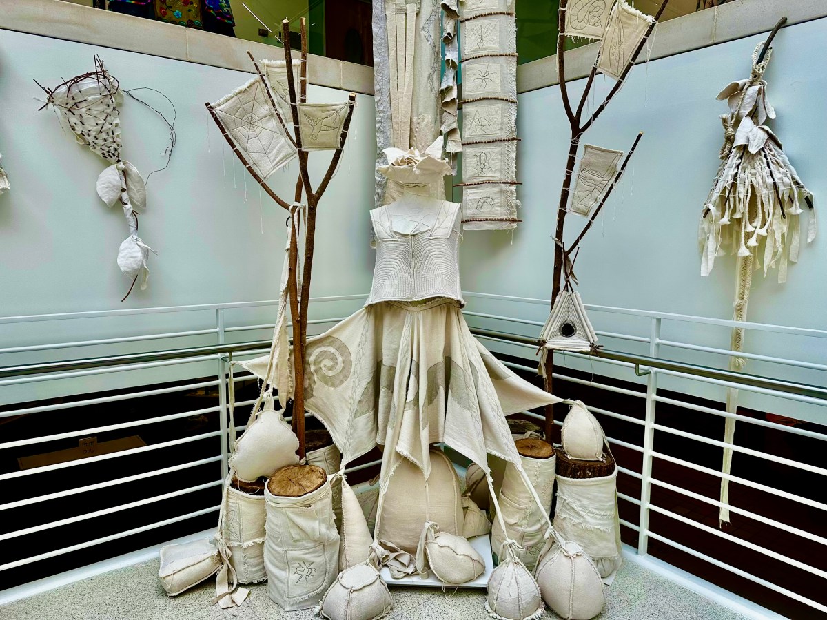 An all-white textile dress installation surrounded by fabric sculptures, embroidered panels, and branch structures at the Embodied exhibit