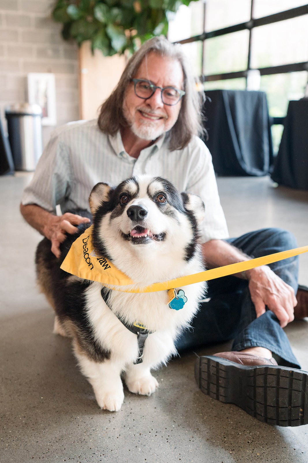 How to get tickets now for the Picasso Pets gala—give back + win a trip to Italy, August 22 2 A smiling man with long gray hair and glasses sits on the floor with a black and white Corgi wearing a yellow bandana reading "Deacon