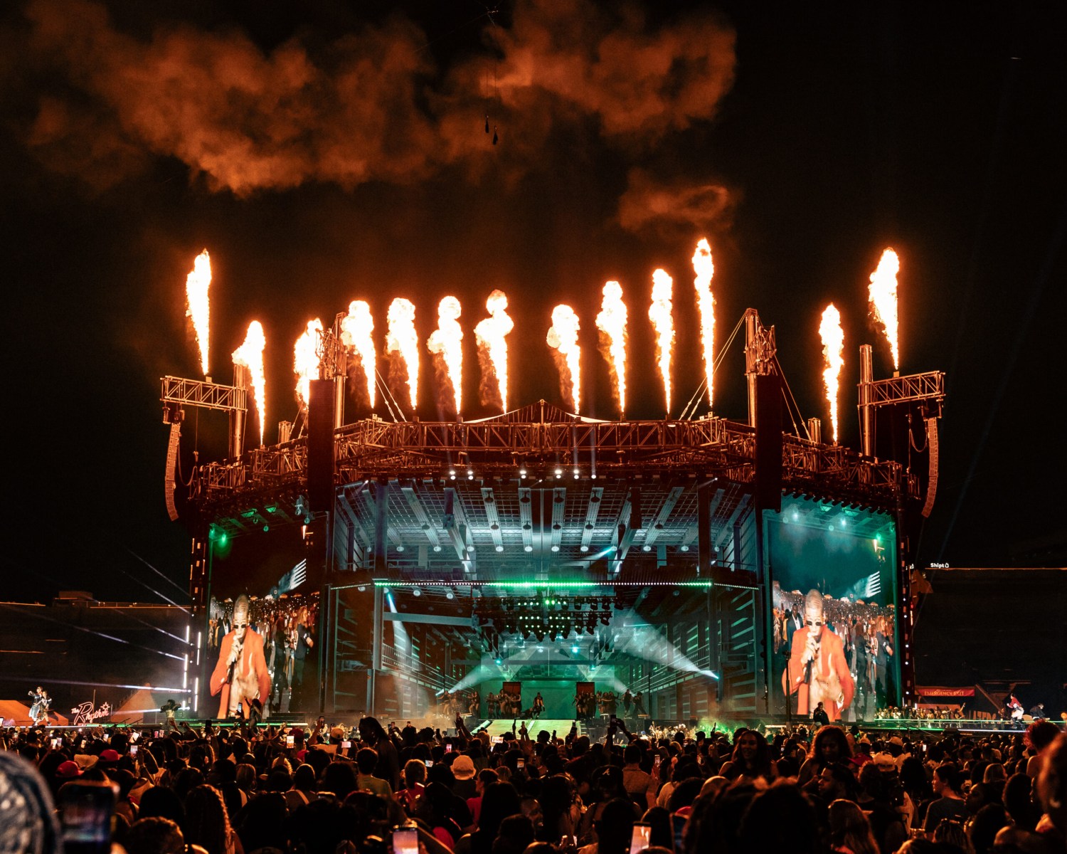 Outdoor concert stage at night with rows of pyrotechnic flames shooting upward, large video screens, and a crowd in the foreground