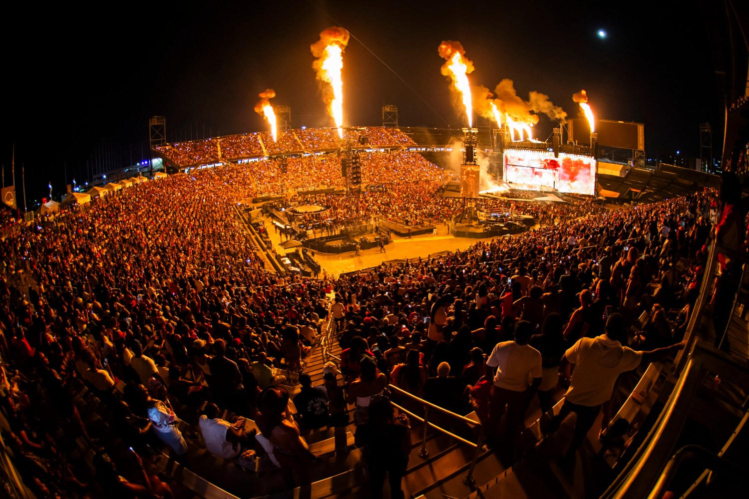 Massive outdoor stadium concert at night with large crowd, stage pyrotechnics shooting tall flames, and a large video screen