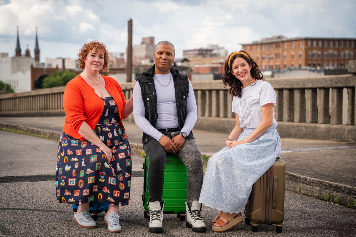Three people posing outdoors on a bridge with luggage, city buildings visible in the background