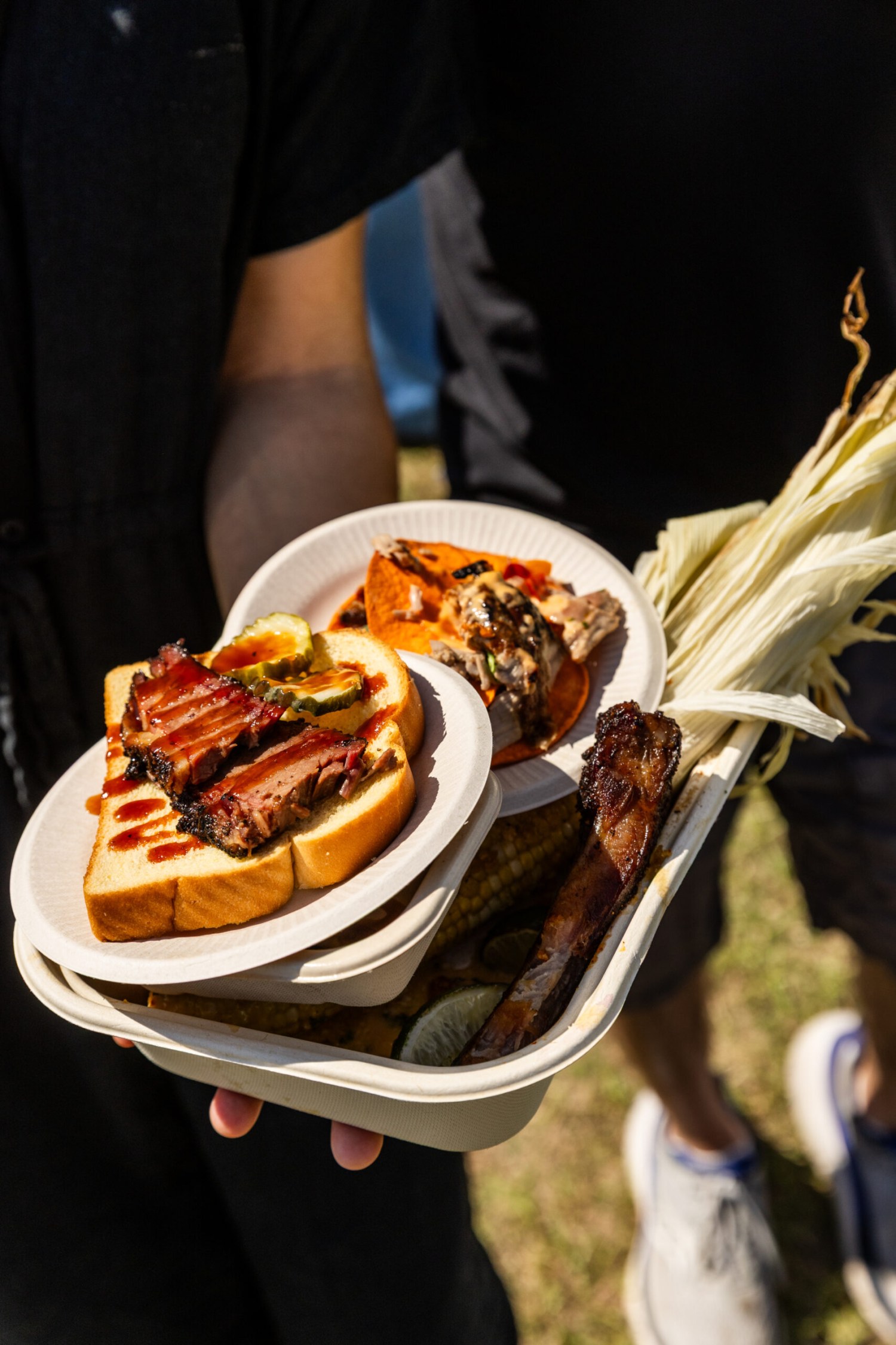 Person holding stacked plates of BBQ brisket sandwich, smoked ribs, corn on the cob, and other barbecue dishes at an outdoor event