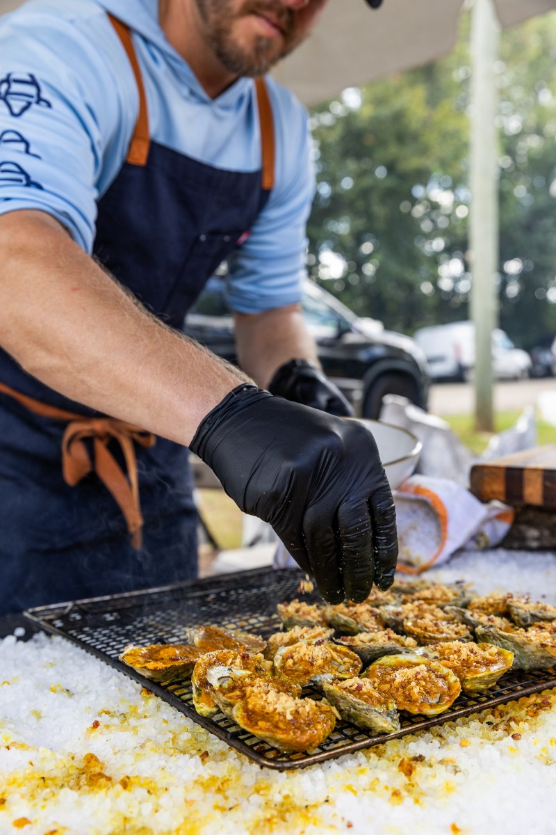A chef in a navy apron and black gloves arranges baked oysters with breadcrumb topping on a rack over crushed ice at an outdoor event