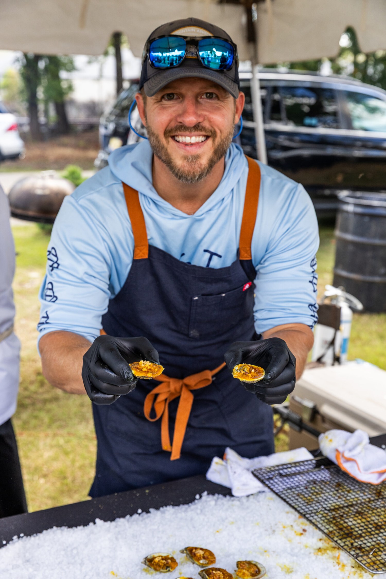 Smiling chef in a navy apron and black gloves holding two topped oysters on the half shell at an outdoor food event