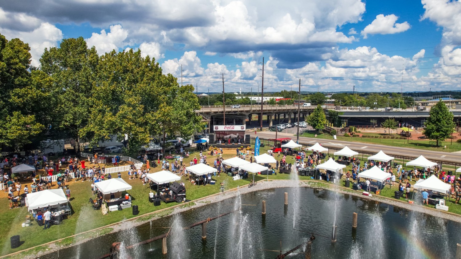 Aerial view of an outdoor festival with white vendor tents, a Coca-Cola sponsored stage, crowds, and a fountain pond in the foreground