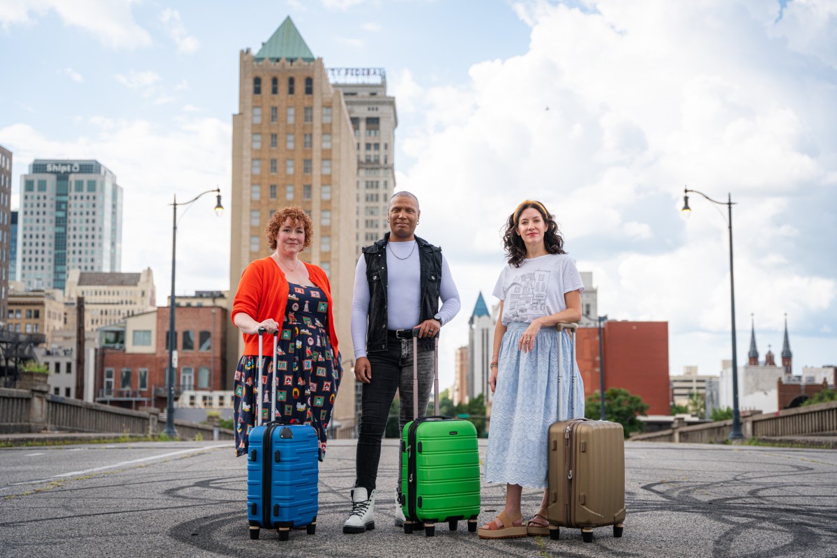 Three travelers standing on a city street, each with a hardshell rolling suitcase in blue, green, and gold, with a downtown skyline behind them