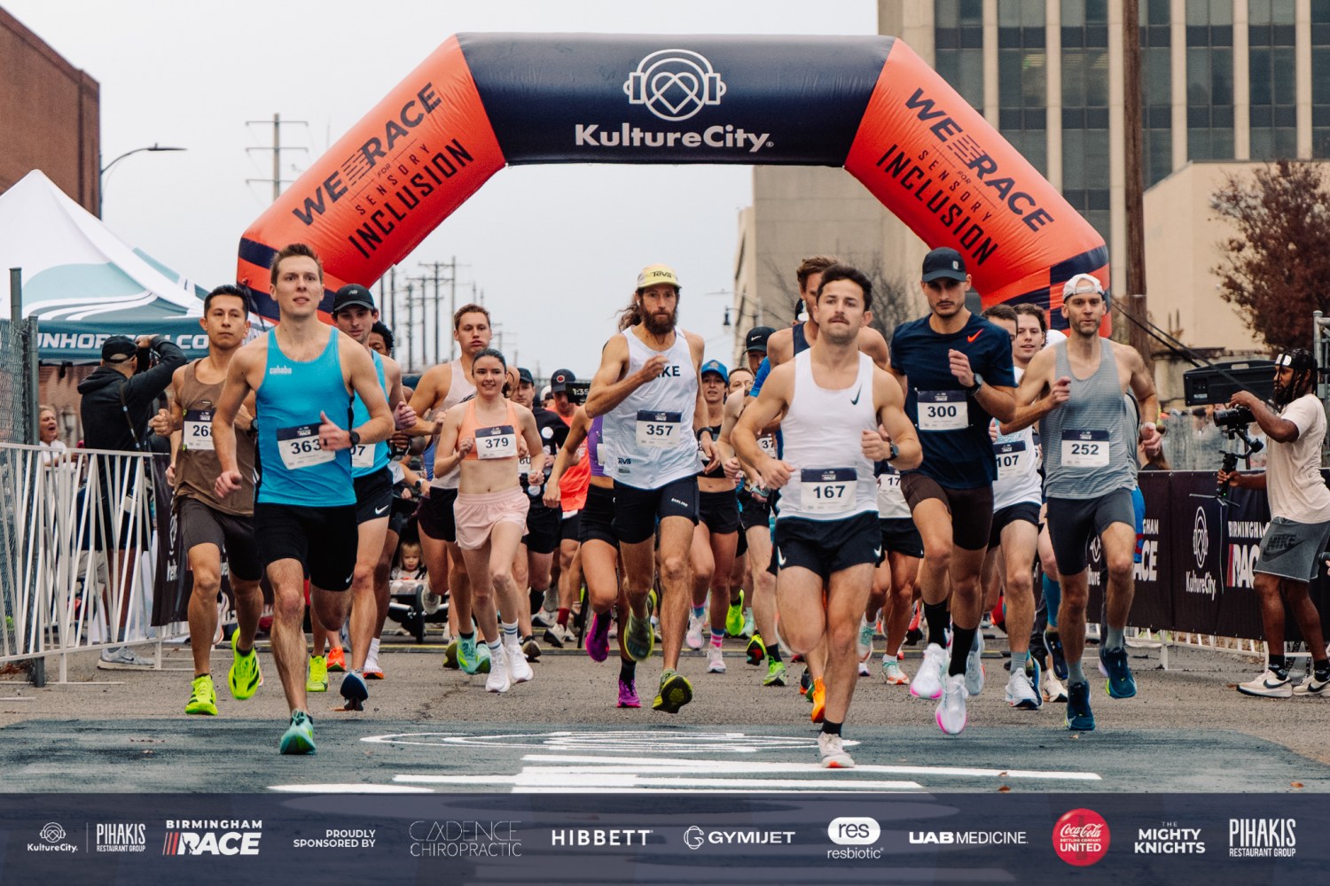 Runners sprint through the start line under a "We Race for Sensory Inclusion" KultureCity arch during a city street race
