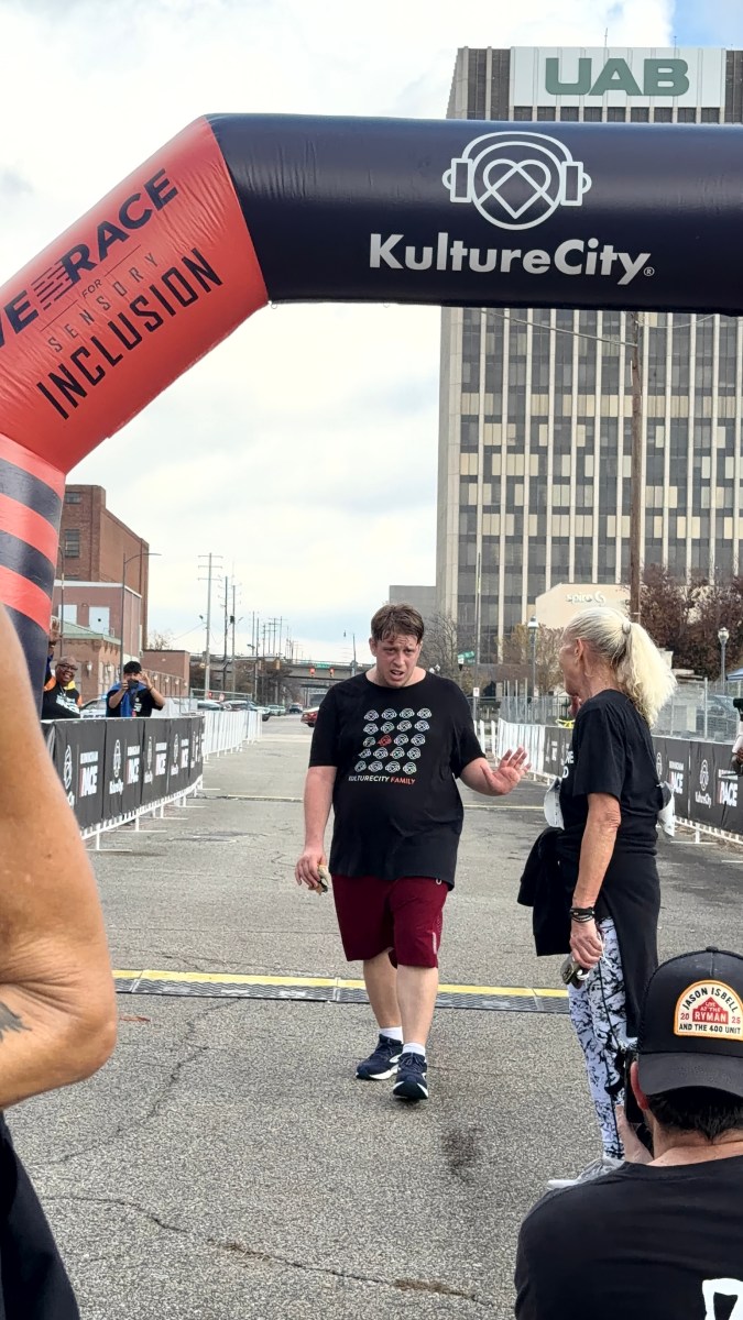 Man in a KultureCity Family shirt crosses under the KultureCity race finish arch on a city street
