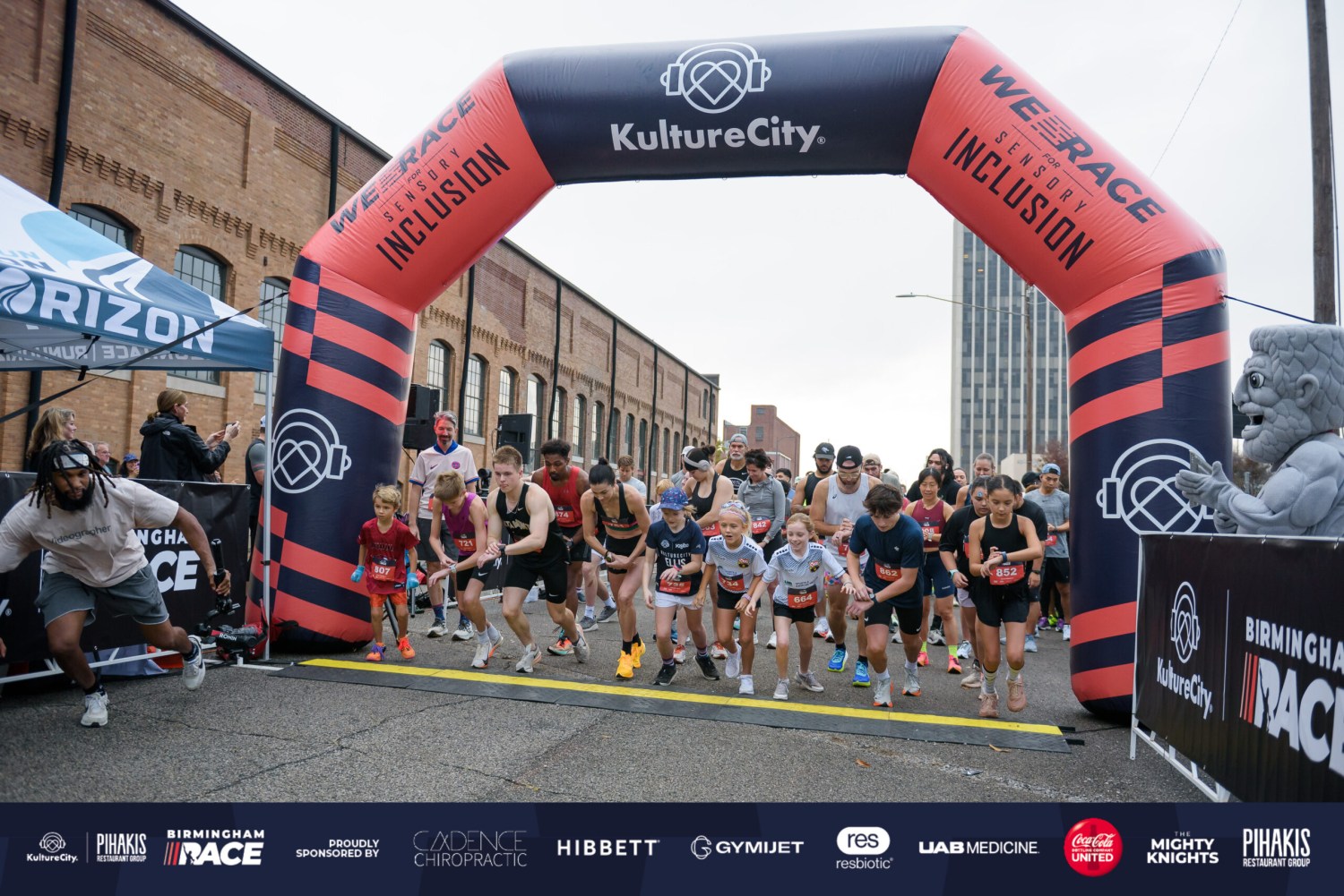 Runners of all ages start a race under a KultureCity "We Race for Sensory Inclusion" inflatable arch on a city street
