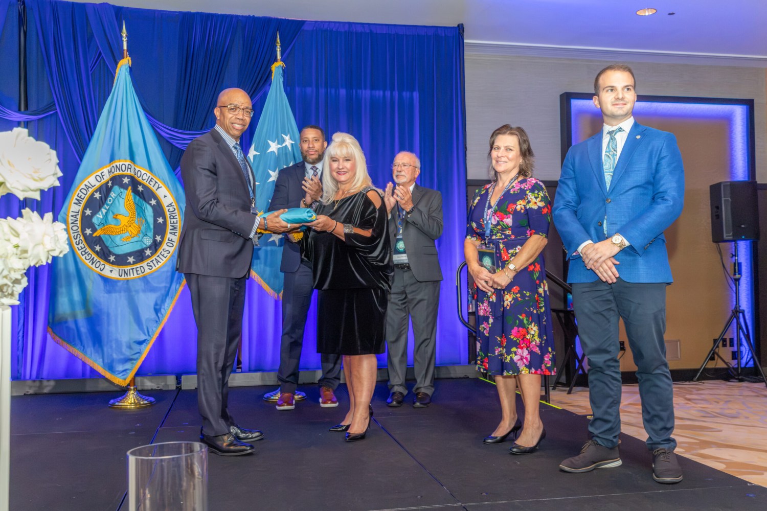 Birmingham recieves Medal of Honor Society Convention flag as 2027 Host City 2 Man presents a wrapped gift to a woman on stage as others applaud, beside the Congressional Medal of Honor Society flag