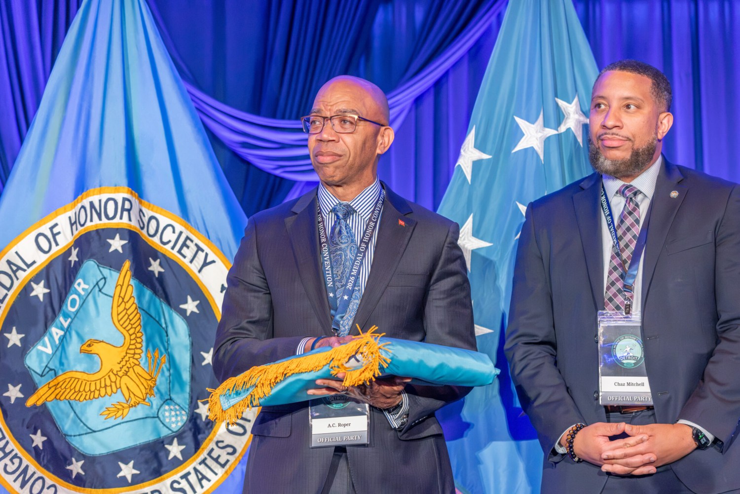 Birmingham recieves Medal of Honor Society Convention flag as 2027 Host City 3 A.C. Roper holds a furled flag beside Chaz Mitchell at the 2026 Medal of Honor Convention, with the Medal of Honor Society seal displayed behind them