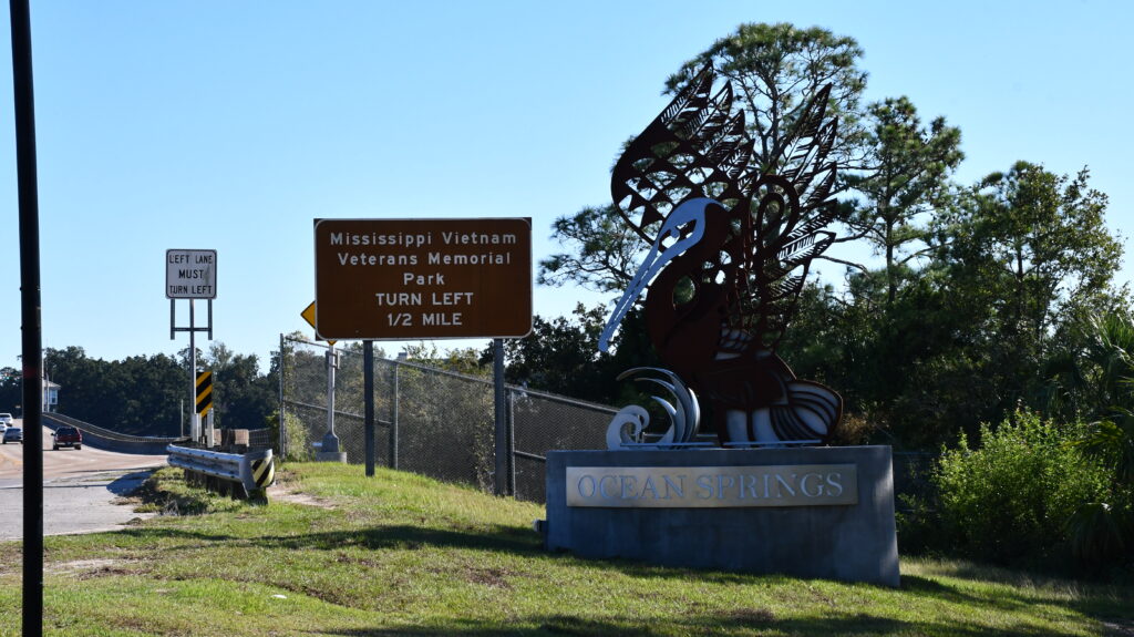 A large metal pelican sculpture stands near a bridge in Ocean Springs, Miss., on a concrete base bearing the city's name.